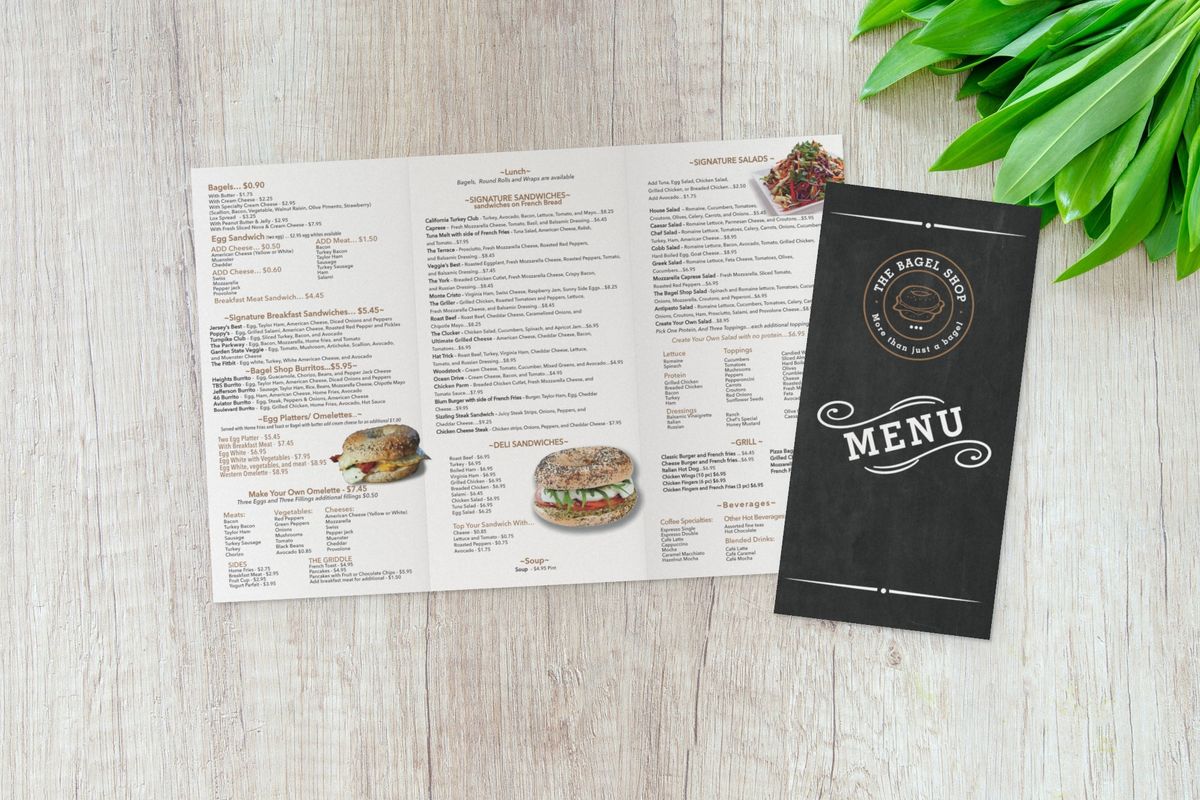 The Bagel Shop tri-fold brochure open to three panels of menu items and prices with food photos, partly covered by its chalkboard-style front panel, on a light wooden surface.