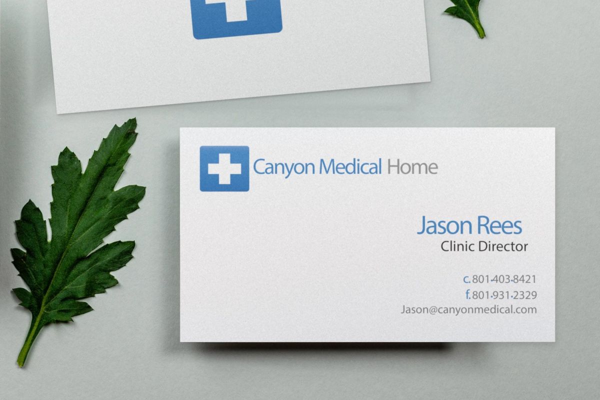 Two Canyon Medical Home business cards on a pale gray-green surface beside two green leaves: one shows a blue square with a white cross; the other shows the logo, Jason Rees, Clinic Director, and phone, fax and email details.