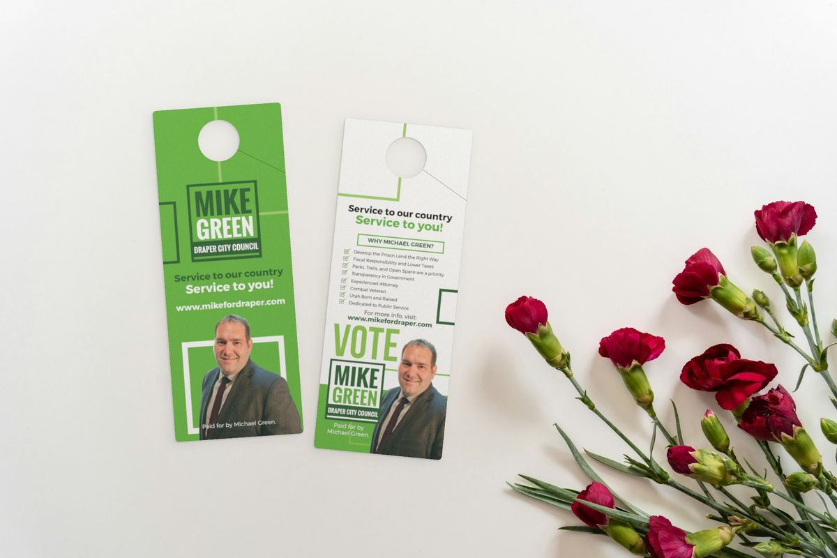 Front and back of a green Mike Green city council campaign door hanger, each with a round die-cut hole and slit at the top, beside red carnations.