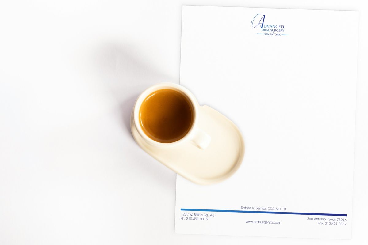 Advanced Oral Surgery of San Antonio letterhead with a blue line-art logo at the top and a blue gradient rule over the contact details at the bottom, beside an espresso cup.