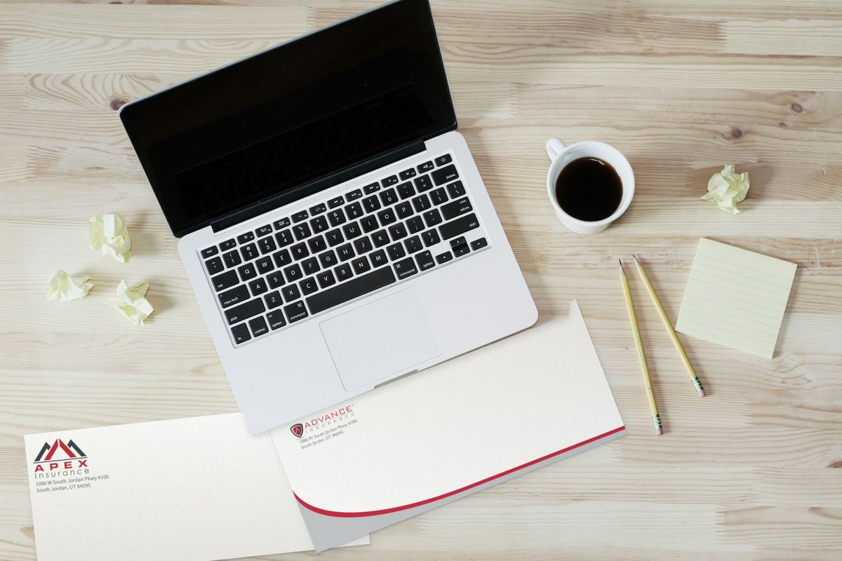 Two business envelopes on a wooden desk below a laptop and coffee: one for Apex Insurance with a red and gray mountain logo, one for Advance Insurance with a red and gray curve along the bottom.