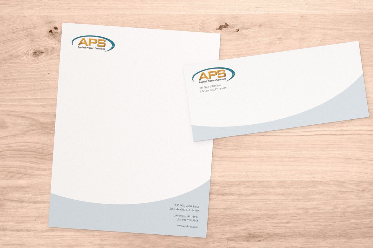 Applied Product Solutions letterhead and matching envelope on a light wood table, each with the gold and teal APS logo and a pale gray curve along the bottom.