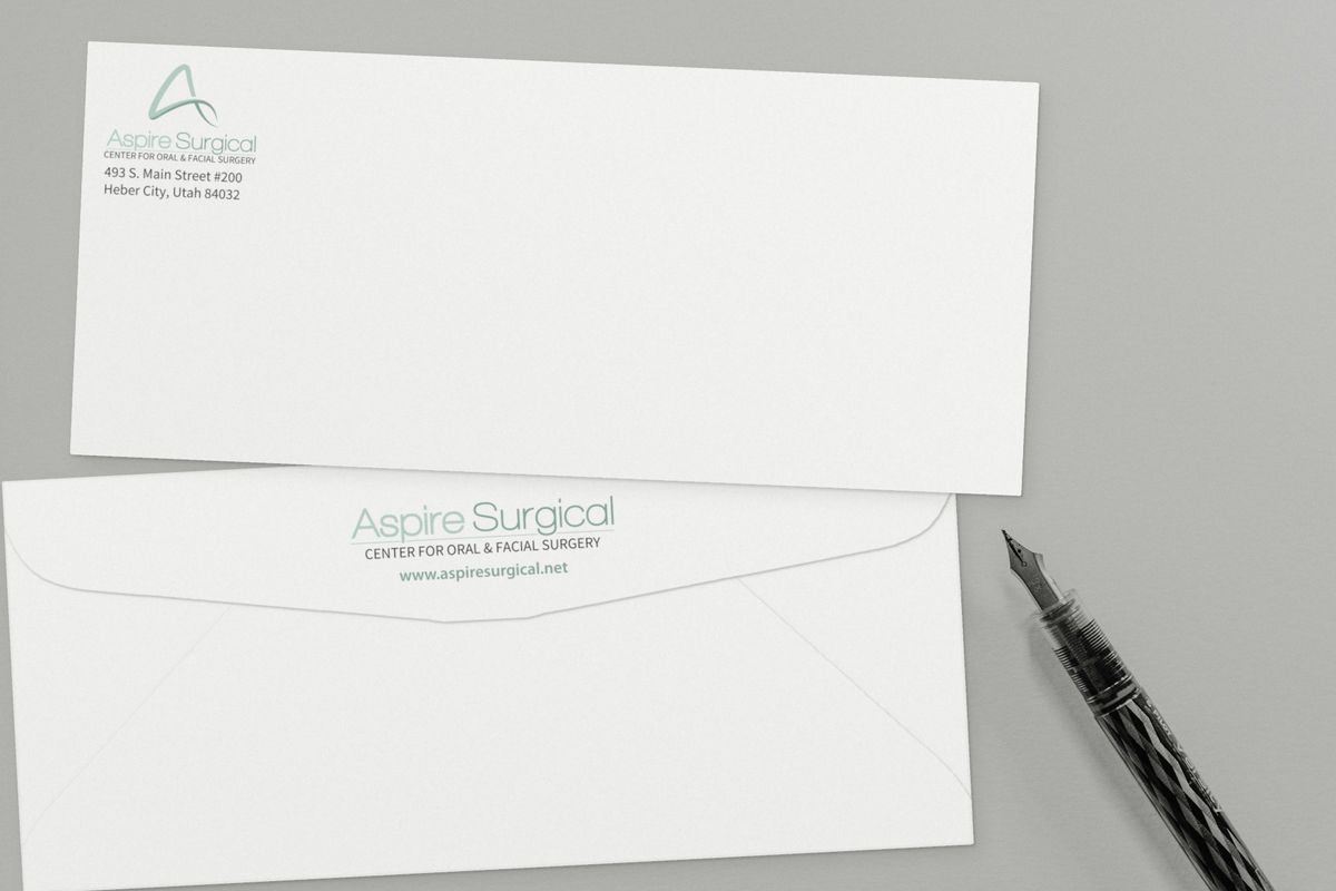 Two white Aspire Surgical business envelopes on a gray surface, one showing the logo and return address on the front, the other the logo and web address on the back flap, beside a fountain pen.