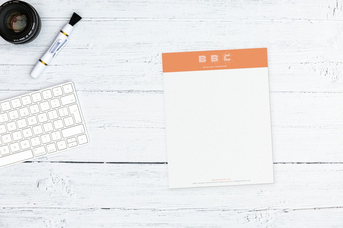 Better Body Compositions letterhead with an orange header band holding the white BBC logo and a one-line contact footer, on a white wooden desk beside a keyboard and camera lens.
